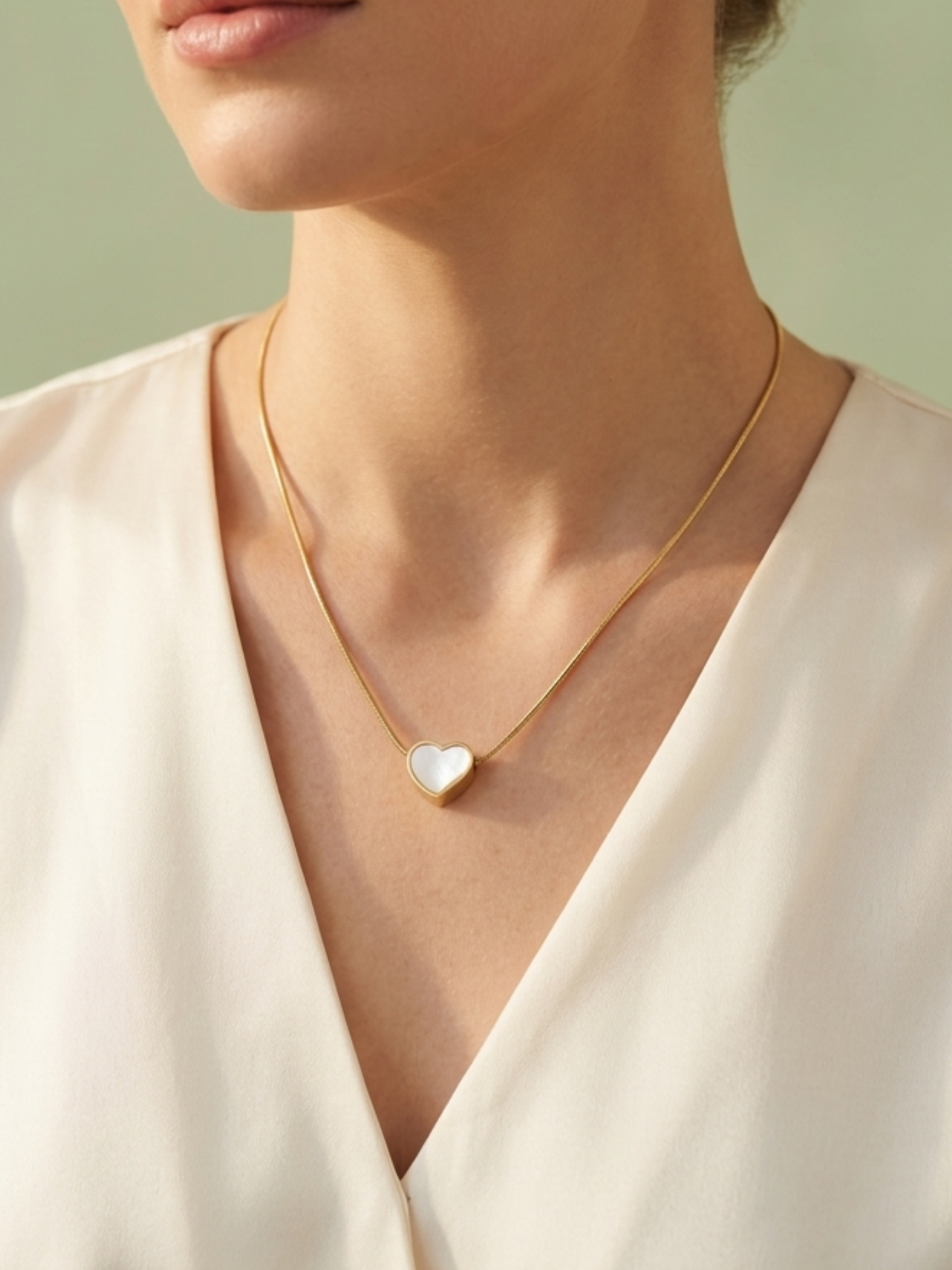 Gold necklace with a heart-shaped pendant worn by a person in a white blouse against a light green background