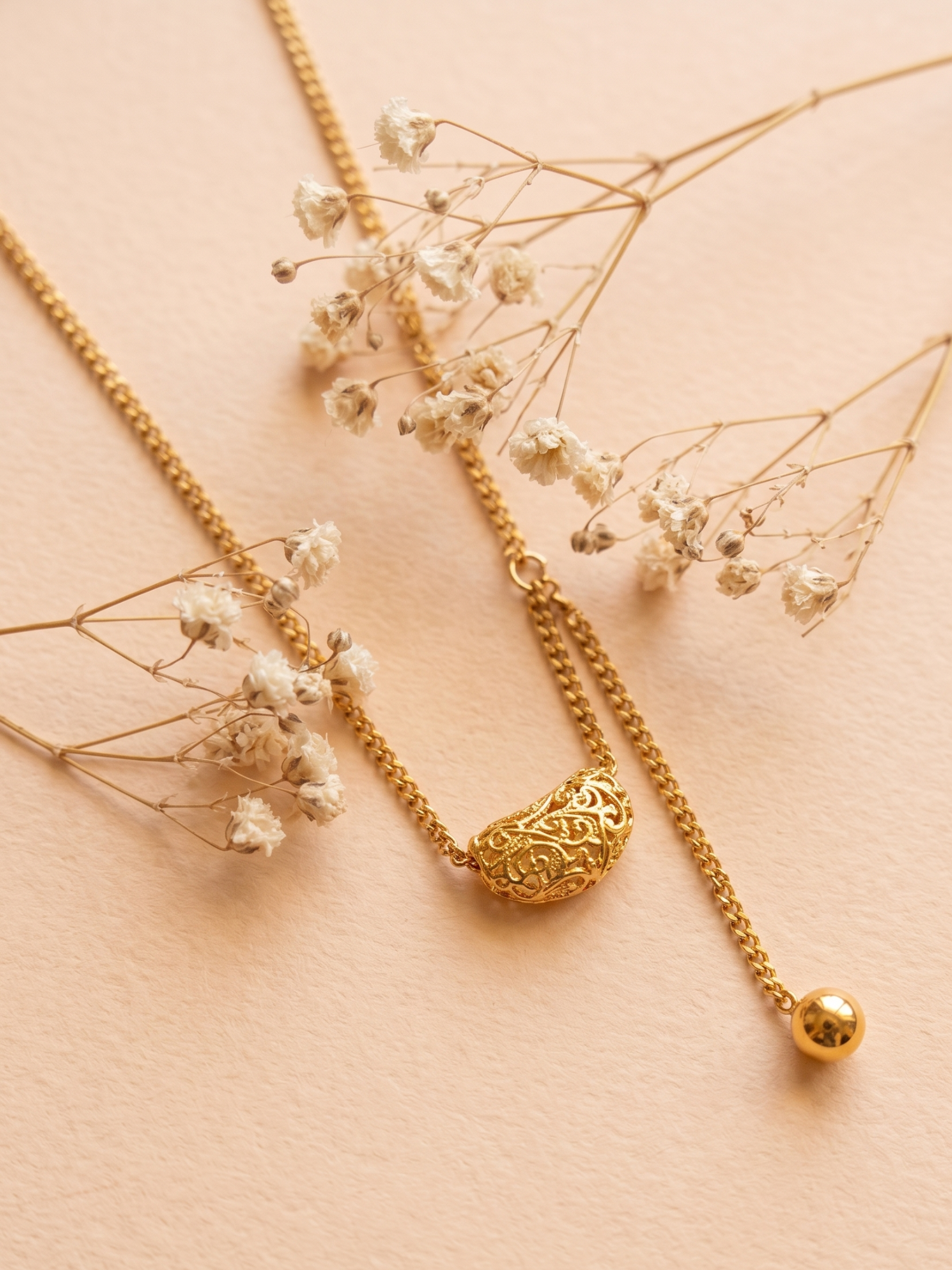 Gold necklace with intricate pendant on a soft pink background with dried flowers
