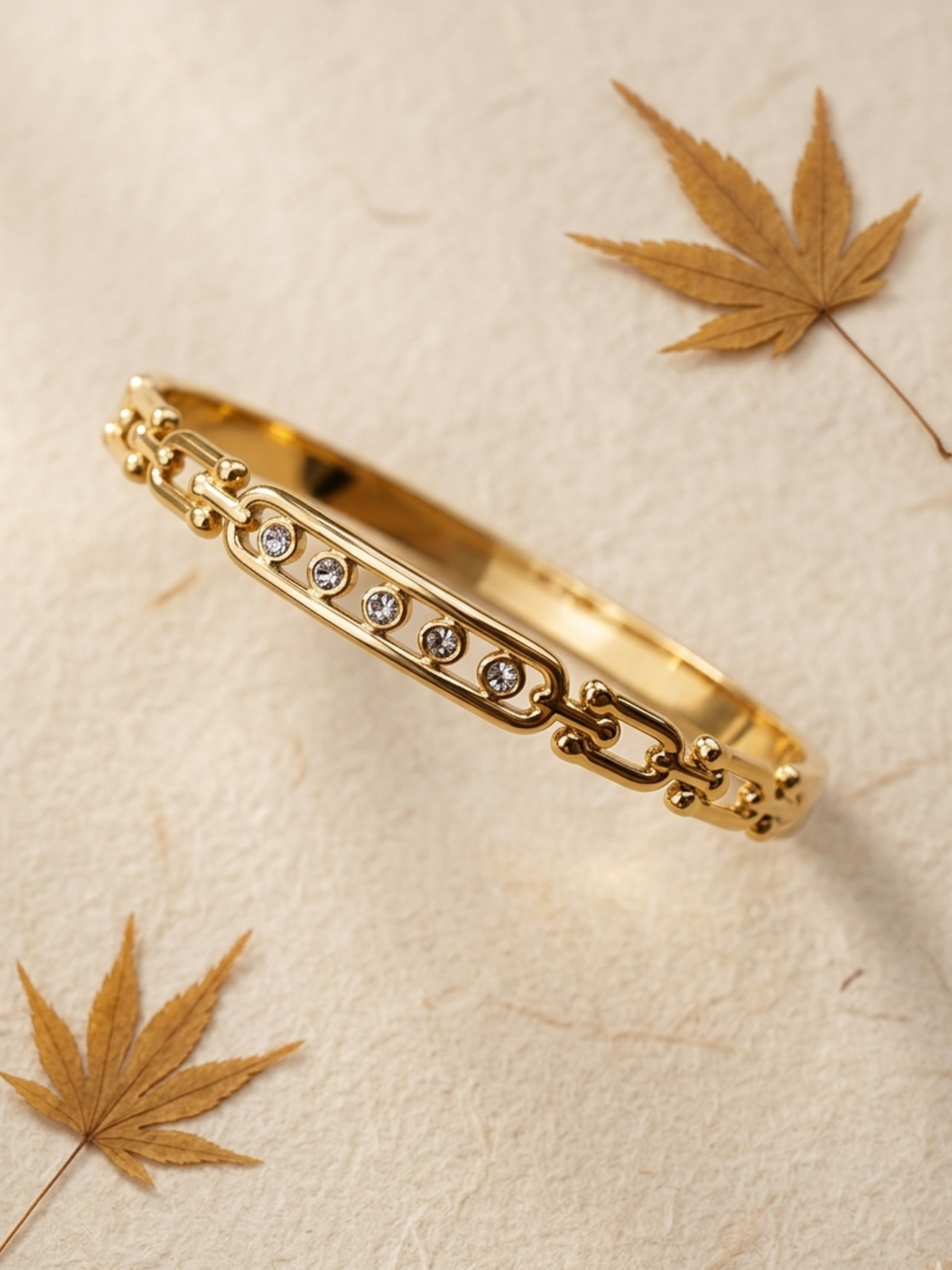 Gold bracelet with embedded stones on a beige surface with leaves